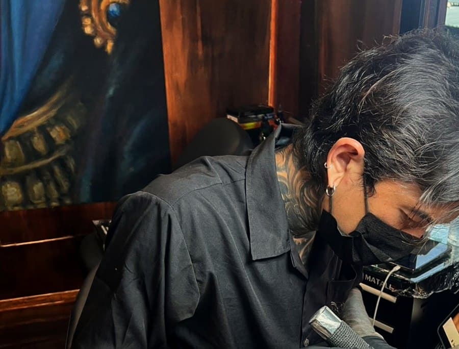 Tom tattooing at 9 House — machine in a gloved hand, working on a client under the painted deity canvas